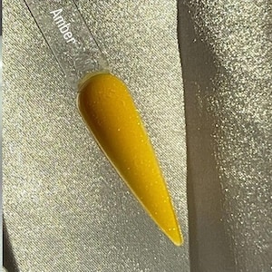 Może przedstawiać: Próbnik paznokcia z żywym, bursztynowym lakierem do paznokci. Przezroczysty plastikowy patyczek jest oznaczony napisem "Amber". Lakier ma błyszczące wykończenie z subtelnymi drobinkami brokatu, tworząc efekt połysku. Tło to teksturowana, srebrna tkanina.