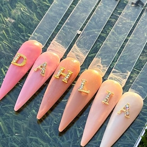 May include: Six artificial nails in shades of pink and peach, each adorned with a gold letter. The nails are arranged on clear plastic sticks. The background is a green surface, likely grass or a similar texture.
