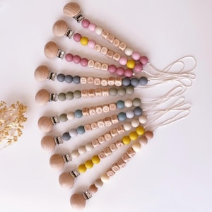 May include: A collection of personalized baby pacifier clips. Each clip features wooden beads, colorful silicone beads, and a wooden clip. The names "NOVA", "ELIZA", "AVA", "SAWYER", "OWEN", "JAXON", "TYLER", "OZZY", and "OSCAR" are spelled out with wooden letter blocks.