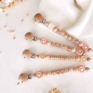 Puede incluir: Cuatro clips de chupete para bebés de madera, con nombres escritos con cuentas de letras de madera. Los clips tienen cuentas de madera, detalles de flores rosas y crema, y ​​soportes de chupete de madera. Los nombres son Millie, Jules, Amelia y Emma.