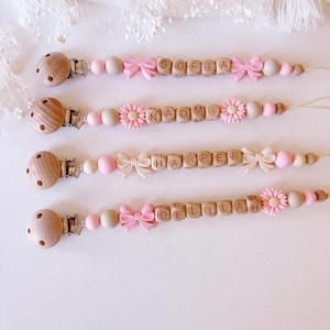 Puede incluir: Cuatro clips para chupete personalizados con cuentas de madera natural, cuentas de silicona rosa claro y detalles decorativos de lazos o flores. Cada clip incluye un cierre de madera redondo y cubos de madera con los nombres Sofia, Naomi, Harper y Delilah.