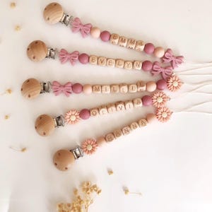 May include: Four personalised baby dummy clips with wooden beads, names spelt out in wooden letter blocks, and decorative bows and flowers. The clips are in shades of pink and peach.