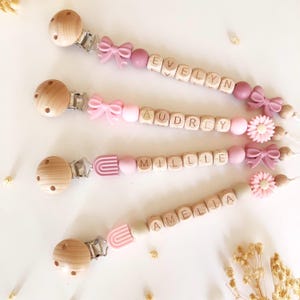 May include: Four baby pacifier clips with wooden beads spelling out names. Each clip has a wooden round clip, a metal clip, and pink accents, including bows and flowers. Names include Evelyn, Audrey, Millie, and Amelia.