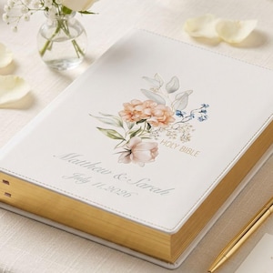 May include: A white Holy Bible with gold gilded edges, featuring a floral design in peach, pink, and blue. The cover is personalized with names and a date. A gold pen and a guest book card are also present.