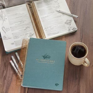 May include: An open teal "My Creative Bible" with gold spiral binding and a closed teal Bible next to it. The open Bible has handwritten notes and illustrations. A cup of coffee, pens, and a green apple are also on the wooden surface.