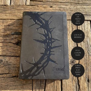 May include: A dark gray journal with a black thorn design and the name "Daniel" embossed on the cover. Additional text on the side indicates the journal has a lay-flat binding, full-color maps, and large print.