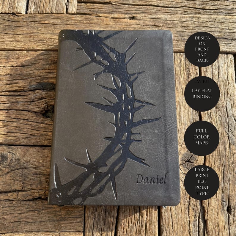 May include: A dark gray journal with a black thorn design and the name "Daniel" embossed on the cover. Additional text on the side indicates the journal has a lay-flat binding, full-color maps, and large print.