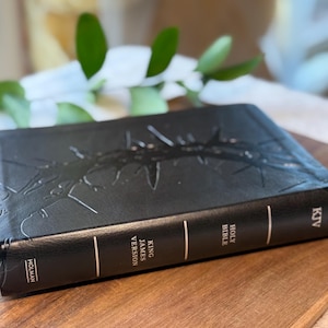 May include: Black leather-bound King James Version Holy Bible with embossed design. The spine features the text "Holy Bible" and "King James Version". The book is on a wooden surface.