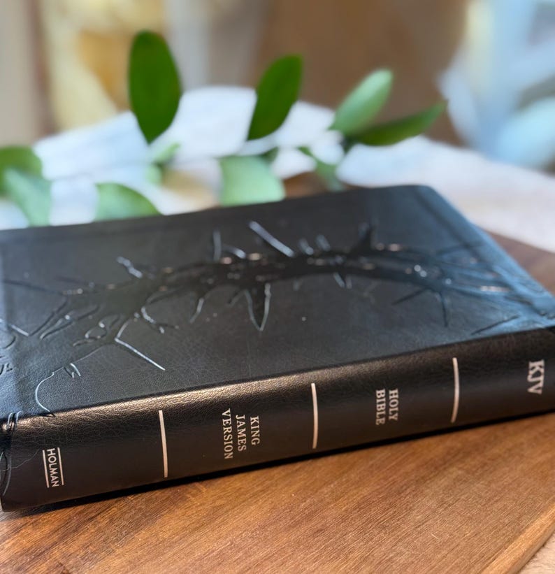 May include: Black leather-bound King James Version Holy Bible with embossed design. The spine features the text "Holy Bible" and "King James Version". The book is on a wooden surface.