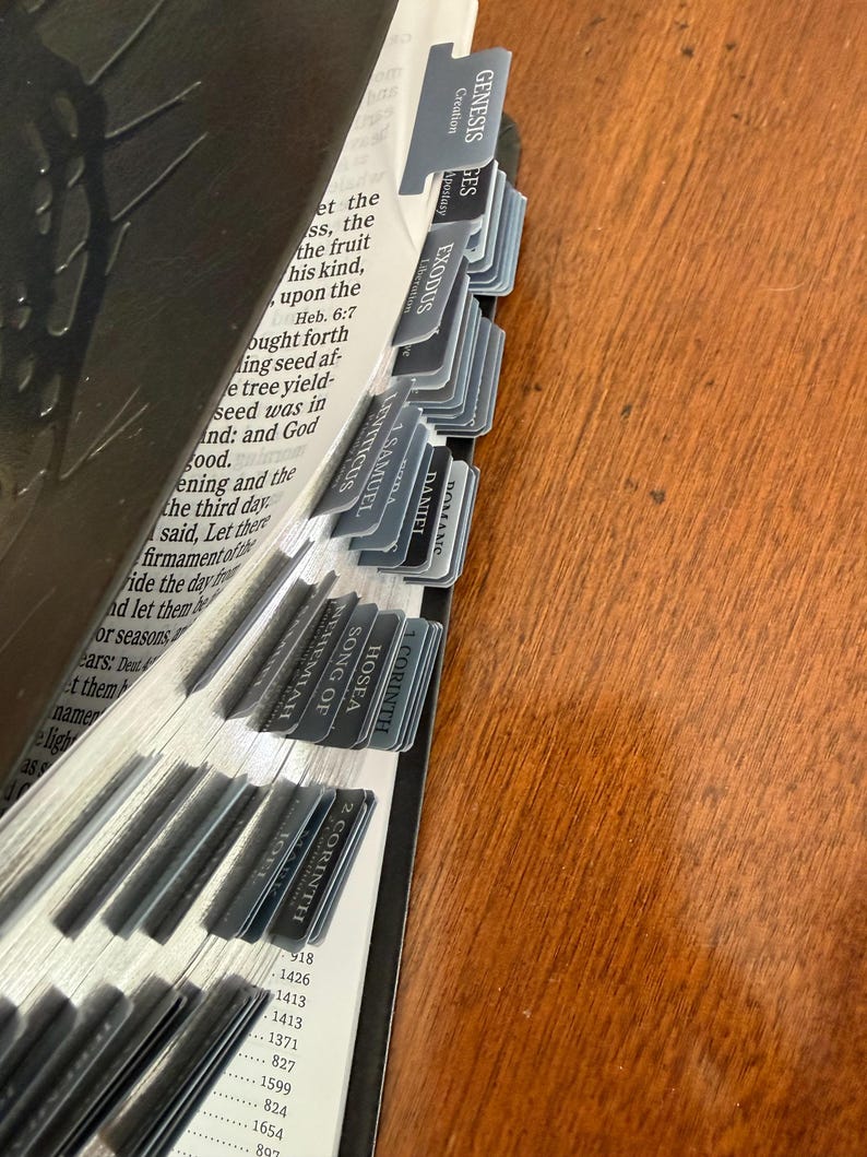 May include: Open Bible with gray tabs labeled with book names like Genesis, Exodus, and 1 Corinthians. The pages are open, revealing text and numbers. The tabs are arranged along the edge of the pages.