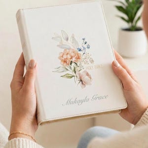 May include: A white Holy Bible with a smooth cover featuring a watercolor floral illustration of peach and blue flowers with green leaves. The text Holy Bible is printed in gold, and the name Makayla Grace is printed in a grey cursive script.