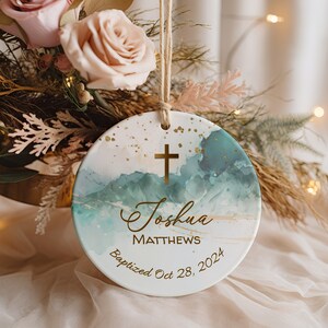 May include: A white ceramic ornament with a blue and green watercolor design and gold accents. The ornament features a gold cross, the name "Joshua Matthews", and the text "Baptized Oct 28, 2024".