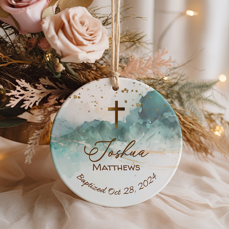May include: A white ceramic ornament with a blue and green watercolor design and gold accents. The ornament features a gold cross, the name "Joshua Matthews", and the text "Baptized Oct 28, 2024".