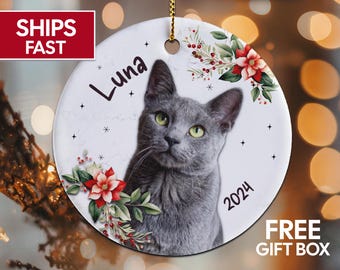 May include: A white ceramic Christmas ornament with a gray cat, red poinsettia flowers, and green leaves. The ornament has the name "Luna" and the year "2024" written on it.