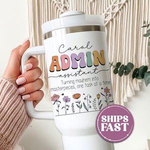 May include: A white stainless steel tumbler with a handle and lid, featuring the text Carol ADMIN assistant and Turning mayhem into masterpieces, one task at a time, decorated with colorful floral illustrations in pink, purple, and orange.