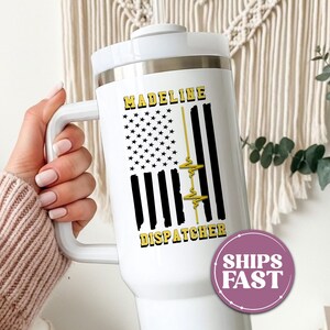 May include: A white stainless steel tumbler with a handle and a lid, featuring a graphic of a black and yellow American flag with a heartbeat line. The name MADELINE is printed in yellow above the flag, and the word DISPATCHER is printed below.