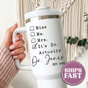 May include: White insulated tumbler with a handle, featuring the text "It's Dr. Actually" and the name "Dr. Jones". The tumbler has a silver band and a white lid. The design includes check boxes for "Miss", "Ms.", and "Mrs.".