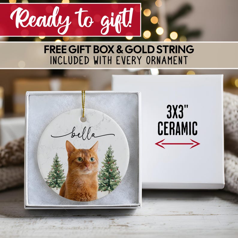K&ouml;nnte beinhalten: Keramik-Ornament mit einer rot getigerten Katze namens Bella, mit zwei gr&uuml;nen Weihnachtsb&auml;umen. Das Ornament ist 7,6 x 7,6 cm gro&szlig; und wird in einer wei&szlig;en Geschenkbox geliefert. Der Text "Ready to gift!" steht oben.