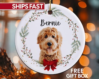 May include: White ceramic ornament featuring a golden doodle dog with the name "Bernie" in black script. The dog is surrounded by a wreath of green leaves and red berries, with a red bow at the bottom. The ornament has a gold string for hanging.