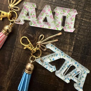May include: Two acrylic keychains featuring Greek letters. One keychain displays the letters Alpha Delta Pi with a pink floral pattern, a gold lobster clasp, and a pink striped tassel. The other shows the same letters with a blue floral pattern.