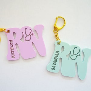 May include: Two personalized "RN" keychains with a stethoscope design. One keychain is light pink with the name "KATELYN" and the other is light blue with the name "SAVANNAH".