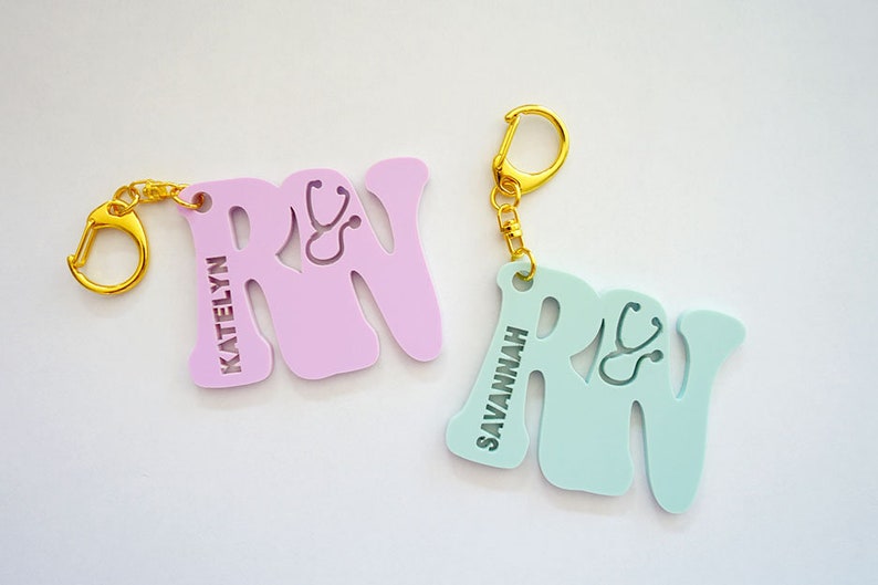 May include: Two personalized "RN" keychains with a stethoscope design. One keychain is light pink with the name "KATELYN" and the other is light blue with the name "SAVANNAH".