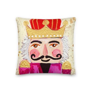 May include: A festive pillow featuring a whimsical painted nutcracker with a gold crown, pink cheeks, and a black mustache. The background is a cream color with gold accents.
