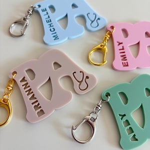 May include: A collection of colorful letter "A" keychains in light blue, pink, and green. Each keychain features a name and a stethoscope symbol. The keychains have silver and gold-colored clasps.