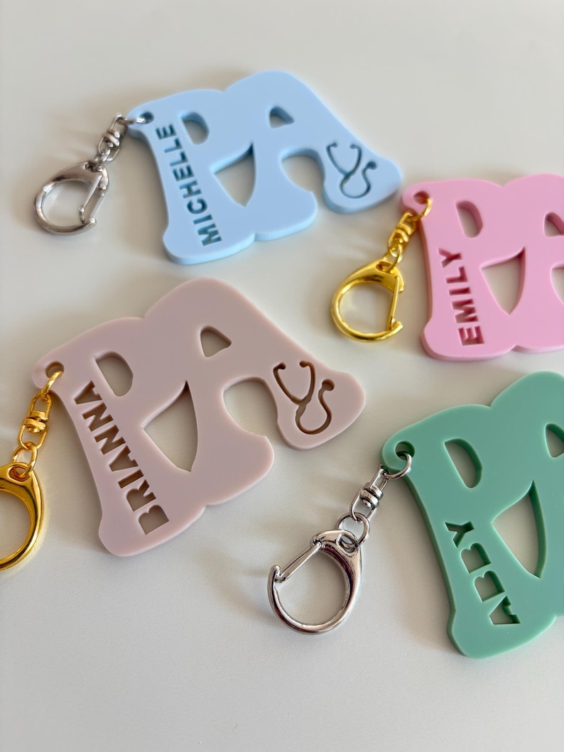 May include: A collection of colorful letter "A" keychains in light blue, pink, and green. Each keychain features a name and a stethoscope symbol. The keychains have silver and gold-colored clasps.