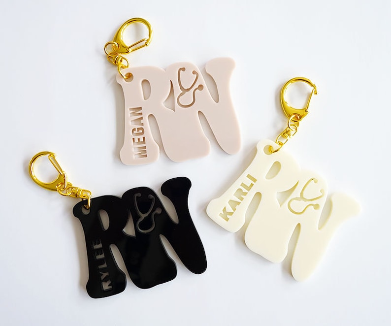 May include: Three personalized keychains with the letters "RN" and a stethoscope symbol. The keychains are black, white, and pink. The names "Kylie", "Karli", and "Megan" are engraved on the keychains.