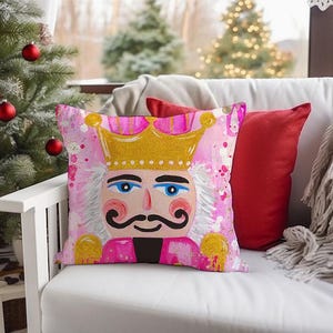 May include: A decorative pillow with a pink background and a painted nutcracker design. The nutcracker has a gold crown, blue eyes, and a black mustache. A red pillow is behind it, and the pillows are on a white couch.