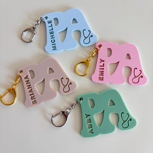 May include: A collection of colorful PA keychains in light blue, pink, green, and mauve. Each keychain features the letters "PA" with a stethoscope design and a name. The keychains have silver and gold-colored clasps.