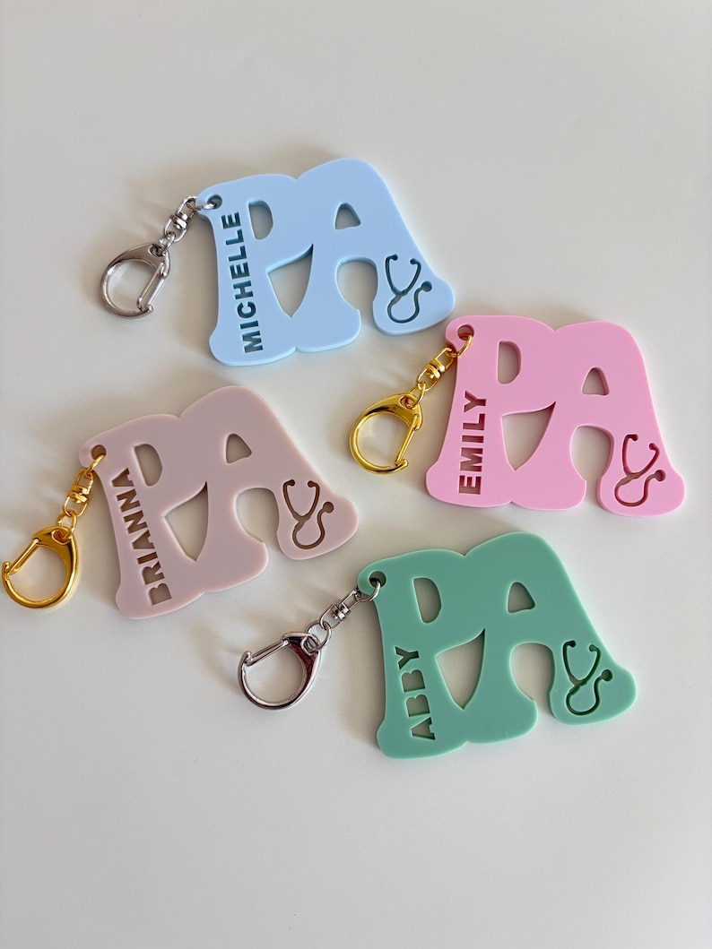 May include: A collection of colorful PA keychains in light blue, pink, green, and mauve. Each keychain features the letters "PA" with a stethoscope design and a name. The keychains have silver and gold-colored clasps.