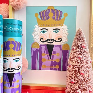 Può includere: Un kit di pittura per numeri con un  cavatappi viola e oro con una corona. Il kit include una tela pre-stampata, barattoli di vernice e pennelli. L'immagine è incorniciata ed esposta accanto a un piccolo albero di Natale artificiale rosa.