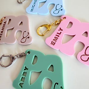 May include: Keychains in pastel colors, each shaped like the letter "A" with a stethoscope design. The keychains are in light blue, pink, and green, with names like "Michelle," "Emily," and "Abby" engraved. Each keychain has a metal clasp.