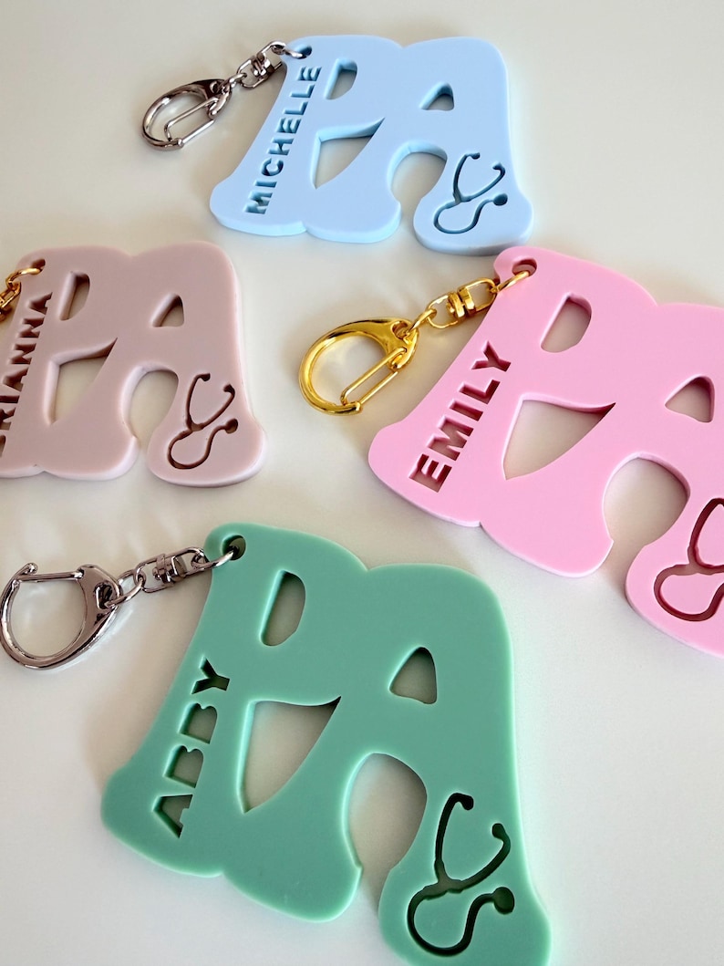 May include: Keychains in pastel colors, each shaped like the letter "A" with a stethoscope design. The keychains are in light blue, pink, and green, with names like "Michelle," "Emily," and "Abby" engraved. Each keychain has a metal clasp.