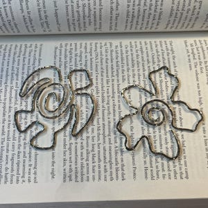 Op de afbeelding: Een zilveren metalen armband met schakels en een decoratief ontwerp. De armband ligt op een pagina van een boek met tekst over een fictieve wereld.