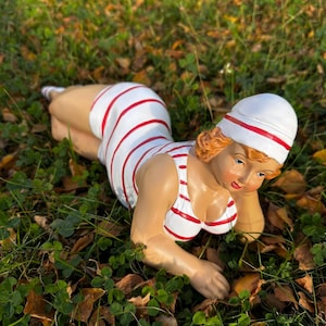 May include: A white and red striped swimsuit with a matching hat. The figure is laying on its stomach with its head turned to the side. The figure is made of ceramic and is a garden decoration.