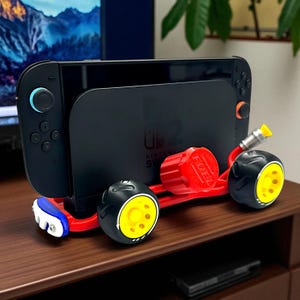 以下が含まれることがあります： 黒いNintendo Switchコンソールが、赤と黒のカート型のスタンドに取り付けられています。スタンドには黄と黒の車輪、赤いエンジン、青と白のアクセントがあります。コンソールは木製の表面にあります。
