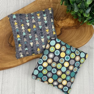 May include: Two small, square fabric items with patterned designs. One features a gray background with rows of yellow, teal, and white triangles. The other has a black background with teal, yellow, and gray circles. The items are on a wooden surface.