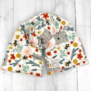 May include: A cream-colored, floral-patterned neck wrap featuring bees, beehives, and flowers in red, yellow, and pink. The wrap is designed to be draped around the neck and shoulders. The fabric has a repeating pattern of bees, flowers, and beehives.
