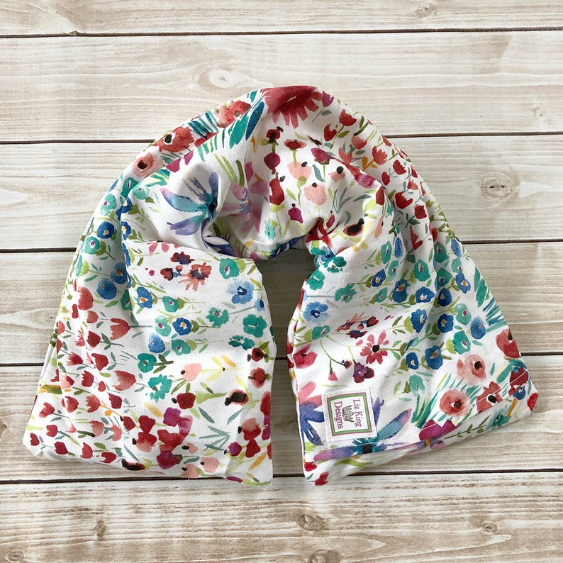 May include: A floral print neck wrap featuring a watercolor design with red, blue, and teal flowers on a white background. The wrap is made of soft fabric and has a label that reads "Lia King Designs".
