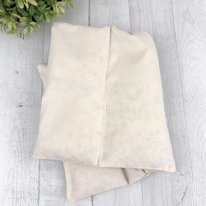 May include: Two off-white, rectangular fabric pouches are stacked on a light wood surface. The pouches appear to be made of a soft, natural material. A small green plant is visible in the upper left corner, adding a touch of color to the neutral scene.