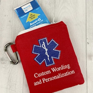 Può includere: Una custodia rossa per emergenze mediche con cerniera bianca e moschettone argentato. La custodia presenta un emblema blu della Stella della Vita e il testo "Custom Wording and Personalization". Un iniettore di epinefrina Auvi-Q è parzialmente visibile.