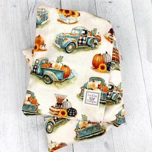 May include: Cream fabric with a repeating pattern of vintage blue trucks laden with pumpkins and sunflowers. The trucks have black and white checkered details. The fabric also features pumpkins, sunflowers, and the text "Crown".