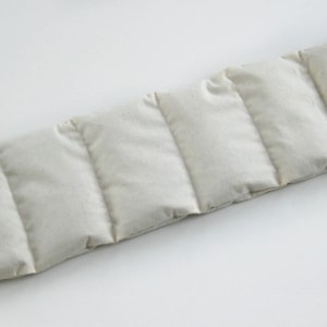 May include: A rectangular, off-white, segmented heat pack. The pack is divided into six sections by vertical stitching. The fabric appears to be a natural, unbleached material. The heat pack is designed to conform to the body.