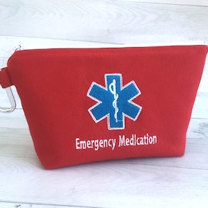Puede incluir: Una bolsa de tela roja con cremallera blanca y mosquetón plateado. Presenta un símbolo médico azul y blanco y el texto "Emergency Medication" en blanco.