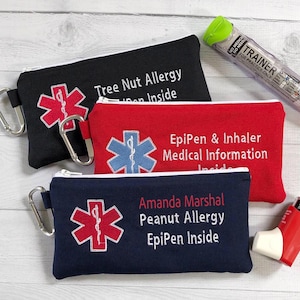May include: Three zipped pouches in black, red, and navy blue, each with a white zip and a silver carabiner clip. The pouches have embroidered medical symbols and text indicating allergy information and EpiPen use. An EpiPen trainer and an inhaler are also visible.
