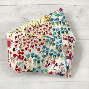 May include: A folded fabric item with a white background, covered in a watercolor floral pattern. The design features red, blue, pink, and green flowers. A small tag with the text "Liz King Designs" is visible.