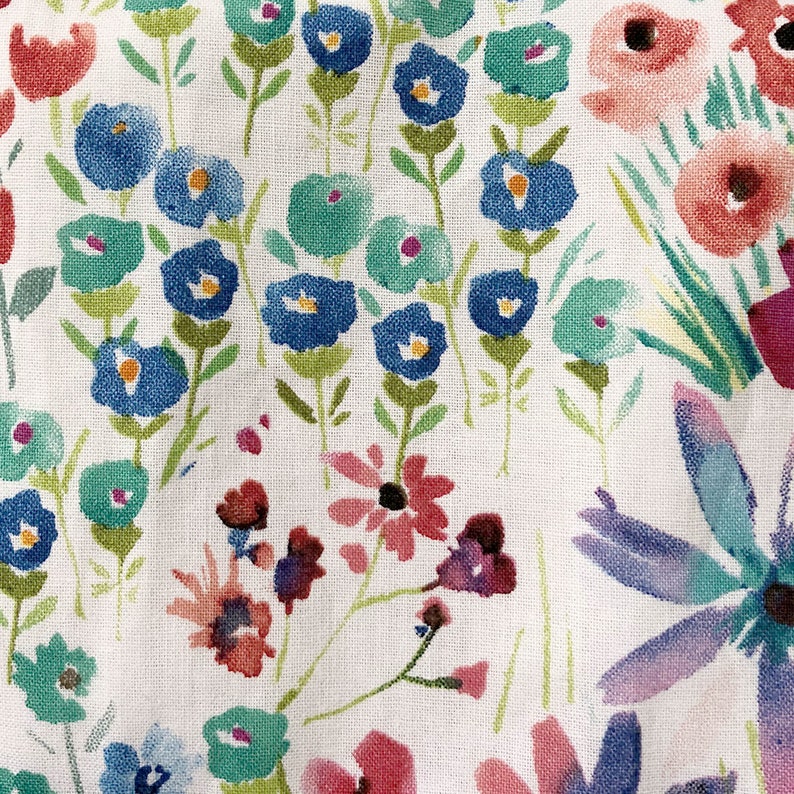 May include: A white fabric with a watercolor floral pattern. The design features blue, teal, red, and purple flowers with green stems and leaves. The flowers are of various sizes and shapes, creating a whimsical look.